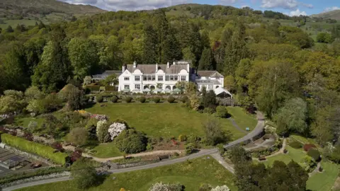 An aerial view of Brockhole-on-Windermere. A large white building is surrounded by gardens and trees.