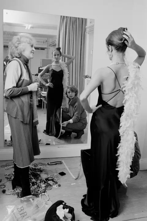 Jean Phenel in a costume fitting, wearing a black backless dress and looking at feather boa in the mirror