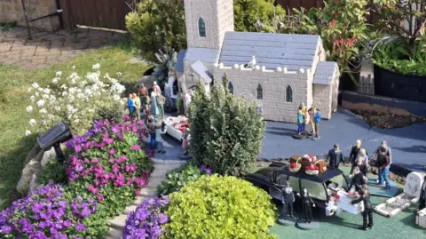 Jane High A miniature church as part of a model village. There is a funeral ceremony playing out with the figures including a coffin being lifted from the back of a hearse. 