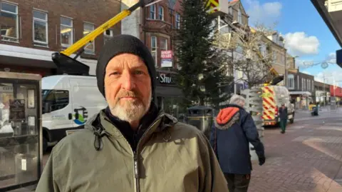 A man with a grey beard wearing a black woollen hat and an olive-coloured coat. He is standing on the street with the tree behind him. Two Denbighshire Council cherry-picker vans are can be seen on either side of the tree.
