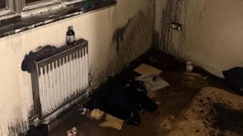 A picture of a soot covered room. There is rubbish on the floor and the outline of where a mattress once was on the carpet. 