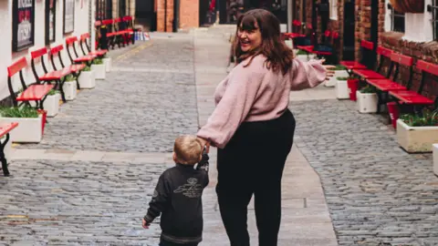 Georgia Anne Photography A woman and a young boy walk hand in hand down a cobbled street. Rows of red chairs can be seen lining each side of the street.