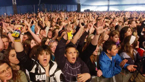A crowd of young people cheer and dance with their arms in the air in an enormous blue marquee. Several are clutching yellow disposable cameras.
