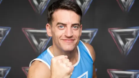 A person wearing a blue and white athletic vest stands with arms crossed in front of a backdrop displaying multiple geometric logos.