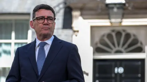 Andy Burnham wearing a dark suit, white shirt and blue tie. He has dark hair and glasses. Burnham is standing outside of 10 Downing Street.