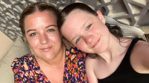 A woman and a girl pose for a picture with their heads tilted to the side. The woman has blonde hair plaited back off her face, she wears a pink, orange and yellow dotted dress and a thin gold necklace. The young girl has dark hair scraped back, and wears a one-strap black top. Behind them both is a detailed silver wall panelling.