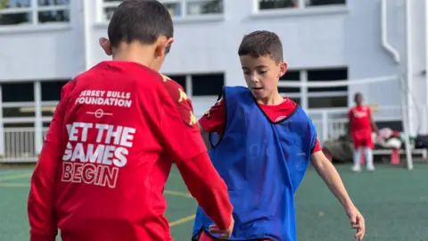 BBC Two boys are playing football. One is wearing a blue bib and is dribbling towards the other boy who has his back to the camera. He has a Jersey Bulls Foundation red top on which has Let the Games Begin on the back. In the background another boy stands in goal. They're playing on an artificial surface at the school playground with a white building behind them.