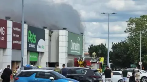 Retail park with smoke billowing from a Dunelm store, flowing over the roofs of neighbouring shops. There is a fire engine by the store entrance and cars parked in the car park. People are standing around watching the fire.