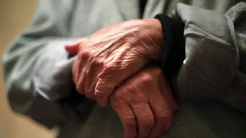 A close up of an old person's hands, they wear a green coat.