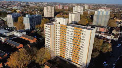 An aerial shot of Barton House in Bristol. It is a large tower block with white and orange panels and lots of windows on it. Other towerblocks and residential properties can be seen in the distance and around Barton House.