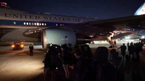 A group of journalists stand on the tarmac next to the Air Force One jet. The white and blue plane stands in the background, with the 'UNITED STATES OF AMERICA' logo visible on the side of the aircraft. The press wait in the dark, dressed in dark colours and checking their mobile phones.