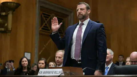 Markwayne Mullin stands, wearing a red patterned tie, a white shirt and a blue suit jacket. He is taking an oath in a wood-paneled room and he is surrounded by sitting people as he prepares to begin his confirmation hearing.