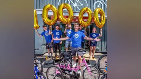 Michael Grisenthwaite Six people, five men and one women, are wearing blue t-shirts with a white logo on them. They are standing behind a blue bike and a pink bike and have large gold helium balloons that spell out the number 10,000