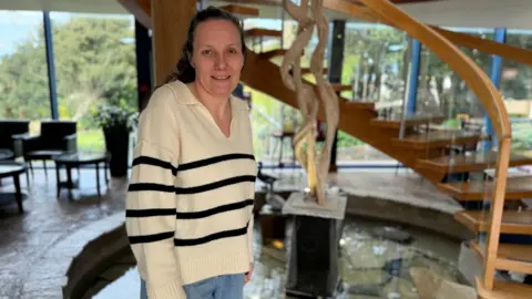 A white women in her mid 30s smiling. Her long brown hair is scraped back off her face. She is wearing a cream v-neck jumper and four thin black stripes going horizontal across. She is standing in front of a water feature which has wooden spiral stairs over it.