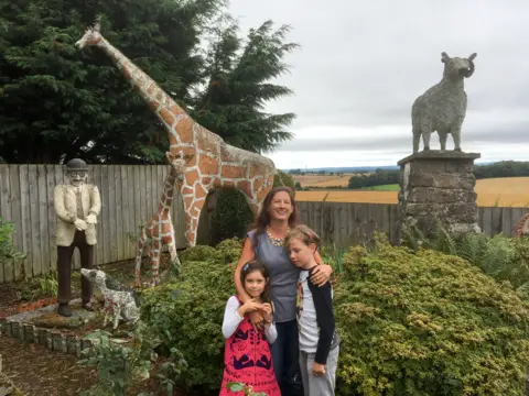 Norma Tattersall A woman with her arms around two children, standing in a rural garden. Behind her are statues of animals including two giraffes. 
