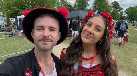 Two people are standing outside in a park wearing traditional Polish outfits.