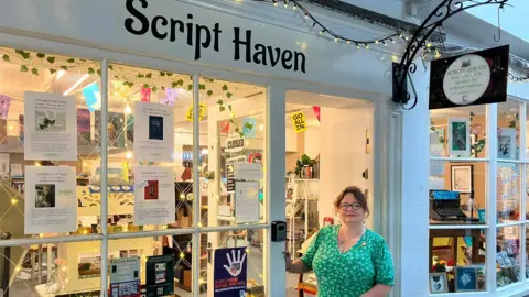Batchelor is stood outside her shop. The front of the store is white with a large shop window. Inside is lit up with warm lighting and several posters are stuck to the window. The shop sign is above the door and reads "Script Haven" in black writing.
