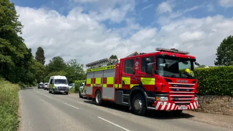 Emergency services at the scene between Leominster and Ludlow, including a fire engine and police van. The vehicles are parked on the road with a hedge behind them.