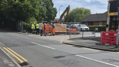 Joy Sinnott A view of construction site, behind a temporary wire fence, with a group of workmen in hard hats and high visibility uniforms gathered in front. 