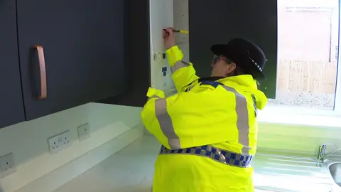 A police officer, wearing a high-vis jacket and hat, marking a boiler with an ultra-violet pen in a kitchen.