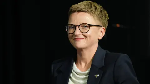 Bev Craig, wearing glasses and a black jacket over a white top, smiles into the camera