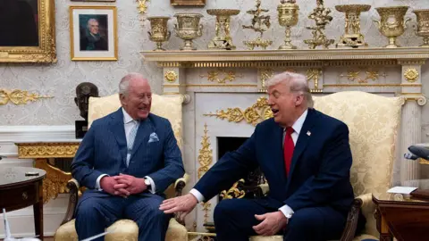 Getty Images King Charles wears a blue pin stripe suit while he sits next to Donald Trump in the Oval Office. Trump wears a dark navy suit and red tie. He touches Charles on the knee whilst the pair laugh. They sit on gold embroided chairs in front of a white and gold decorated wall.
