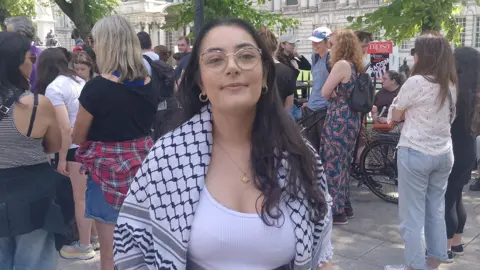 A woman with long brown hair, looking at the camera. She is wearing a white top and a black and white keffiyeh draped over her shoulders.