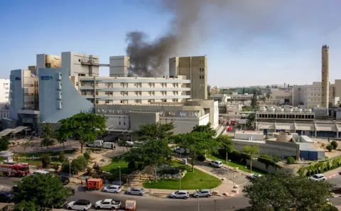 AFP A wide view shows smoke rising from behind the Soroka medical centre against a blue sky and with cars and fire trucks outside it, in Beersheba on Thursday
