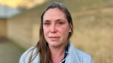 Sara is wearing a grey jacket over a black and white top. She has long, light brown hair and looks serious in her expression.