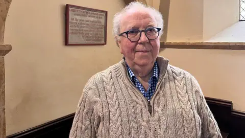 Man with white hair and glasses wearing a blue checked shirt and oatmeal coloured jumper.