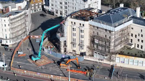 An aerial view of what is left of the Royal Albion Hotel. There is scaffolding around the cream building and three cranes on the road. 