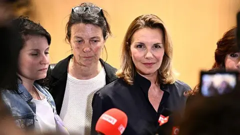 AFP A woman in a dark jacket and blonde hair talks to reporters, with a red microphone in the foreground