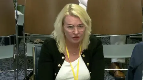 A screengrab of a woman with blonde hair wearing glasses, a white top, black coat and a yellow lanyard sitting at a committee meeting.