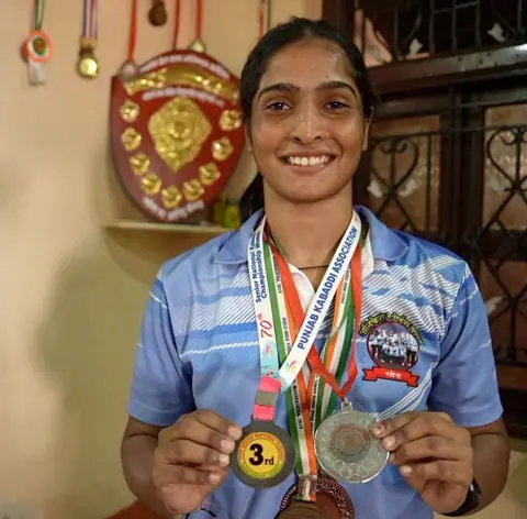 Soves a lot of medals around your neck and more medals in a trophy and background. She is smiling and wearing a light blue sports shirt.