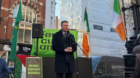 John Finnucane outside court, speaking to crowds from a small stage. He has short dark hair and is wearing a dark knee-length woollen coat over a dark suit, purple tie and white shirt. He also has a navy and dark green tartan woollen scarf. On the stage there are Irish tricolour flags on poles and behind him a red brick building can be seen and also a multi-storey building covered in construction scaffolding and white plastic.