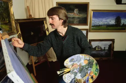 Getty Images A man with brown hair and moustache painting a canvas in a studio. He is wearing a black and grey jumper with a zig zag pattern and a polo shirt. Other paingings are hung on the wall behind him.