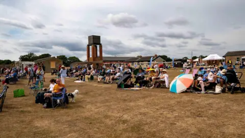 People sitting on grass that looks quite yellow. They have deck chairs and umbrellas. There are about 100 people in the shot.