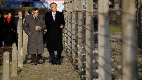 Getty Images Ban Ki-moon, a tall man with short black hair and a long black coat, walks beside Marian Turski, a shorter man in a grey coat with grey hair. They are walking down a path alongside a tall, barbed fence.