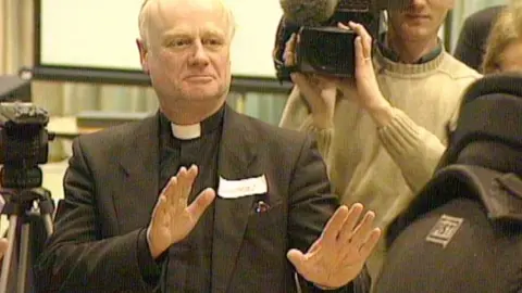 An older man wearing a clerical collar and dark suit raises both hands, palms out, as if signaling “stop,” while cameras and people surround him in an indoor setting.
