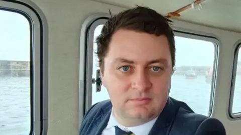 Picture of a man in his mid 30s, with brown hair wearing a suit, on a boat