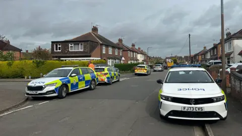 Police in Copeland Avenue near Tevery Close. A number of police cars are parked at the side of the roads in a residential area, two cars are blocking access to Tevery Close and a fire service vehicle can be seen in the background of the image.