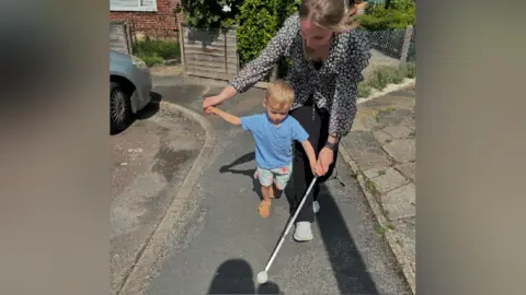Stewarts A young boy wearing shorts and a blue t-shirt, walking with a metal stick in his left hand. He is walking along a pavement helped by a woman behind him.