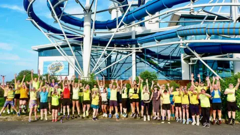 The Southport Strollers A large group of runners dressed in yellow t-shirts are lined up in front of the Southport Splash World water park with their arms in the air.