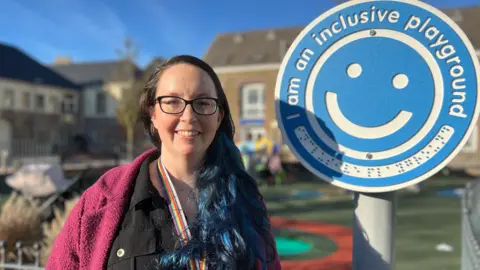 Louise is standing next to a blue circular sign attached to a pole. The sign features a smiling face and the text “I am an inclusive playground.” Louise is wearing a dark outfit with a bright pink outer garment and a rainbow-colored lanyard. Behind her is a fenced playground with colorful play surfaces and buildings in the background.