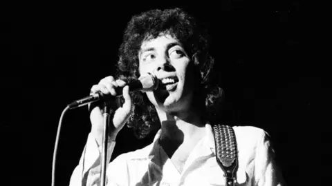Getty Images A black-and-white image of Graham Gouldman on stage in the 1970s, singing into a microphone. He has long curly hair and is wearing a shirt with a guitar strap over his shoulder.