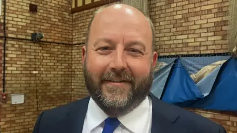 A man in a dark blue suit jacket and blue tie , with a trimmed beard looks at the camera, smiling.