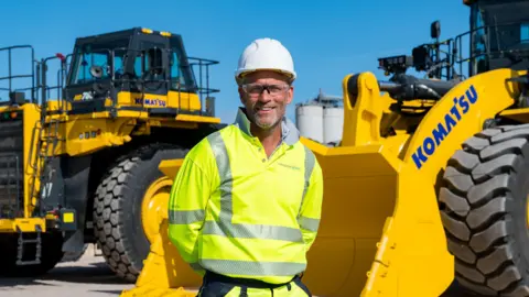 Tungsten West Jeff Court smiling at the camera with a yellow hi-vis. He's stood in front of a large digger.
