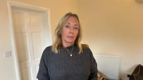 Tracy Baker with long blond hair wearing a grey top and a silver necklace. She is leaning back on a settee covered in a green throw.