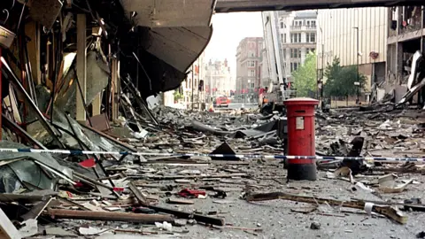 Scenes of devastation following the IRA bomb in Manchester city centre on 15 June 1996. Rubble and debris cover the streets while a red postbox stands apparently unscathed.