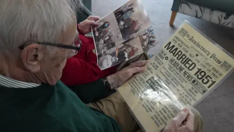Tony and Joy look at old photographs and a newspaper-style anniversary card. . Tony is an older man with short white hair, and glasses. He wears a dark green jumper. Joy is an older woman, with short white hair. She wears a red blouse.